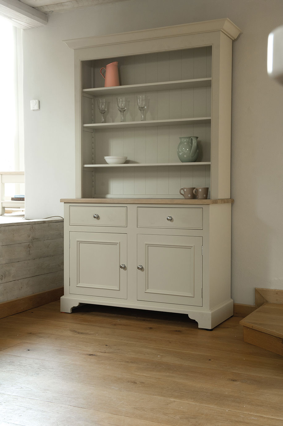 Buffetschrank im Landhausstil aus Holz, cremefarben, mit offenen Regalen, dekoriert mit Gläsern, Krügen und Tassen, in einem hellen Raum auf Holzboden