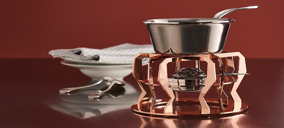 Fondue-Set mit glänzendem Kupfergestell, Edelstahltopf und Brenner, im Hintergrund Teller, Servietten und Besteck auf einem Tisch vor rotem Hintergrund