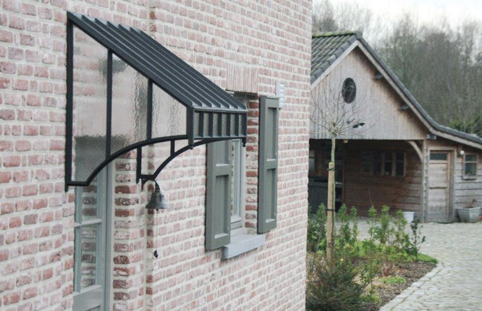 Vordach aus Glas und Metall an einer Hauswand mit Backsteinfassade und Fensterläden, im Hintergrund steht ein Holzhaus