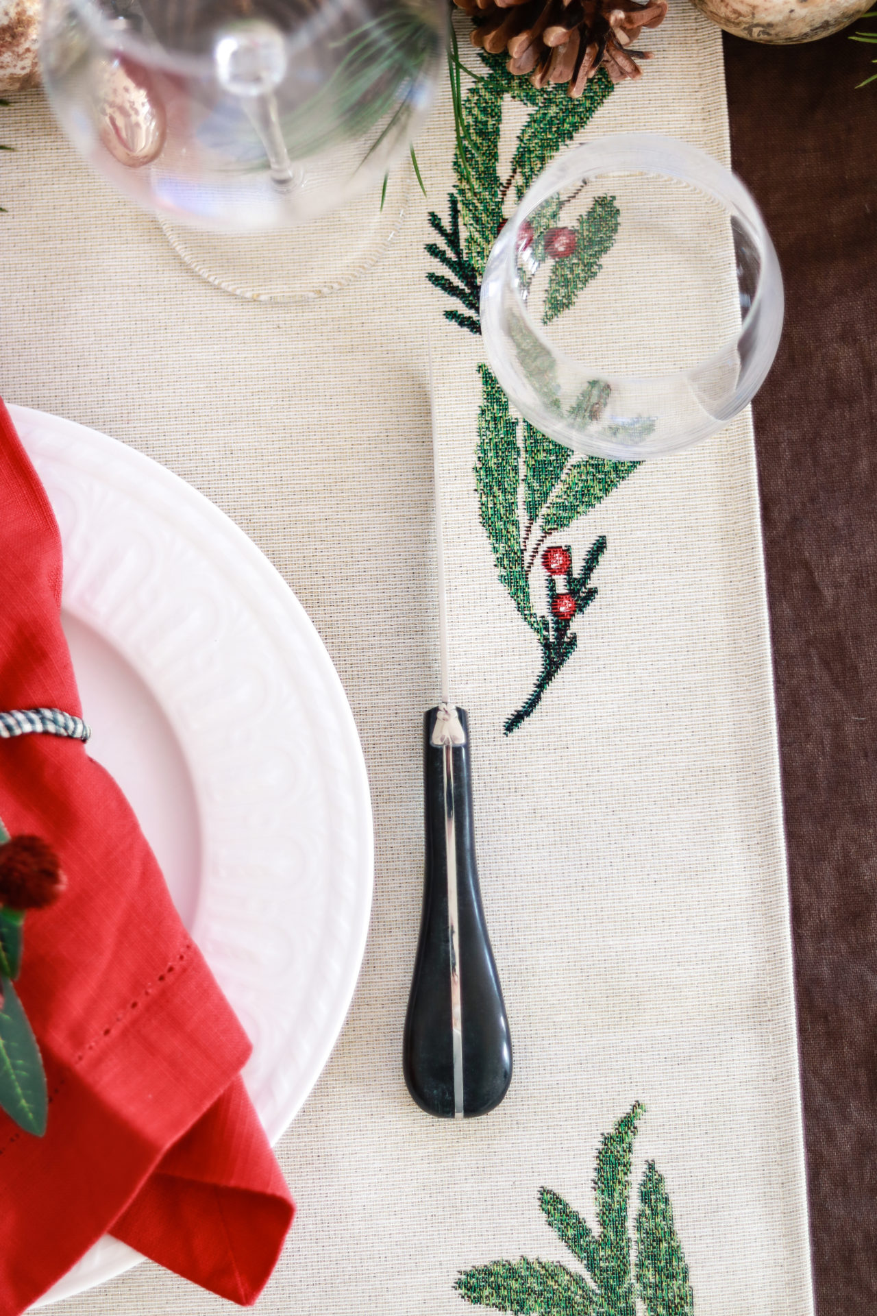 Nahaufnahme eines festlich gedeckten Tisches mit weißem Teller, roter Stoffserviette, schwarzem Besteck und weihnachtlich besticktem Tischläufer mit grünem Zweig und roten Beeren