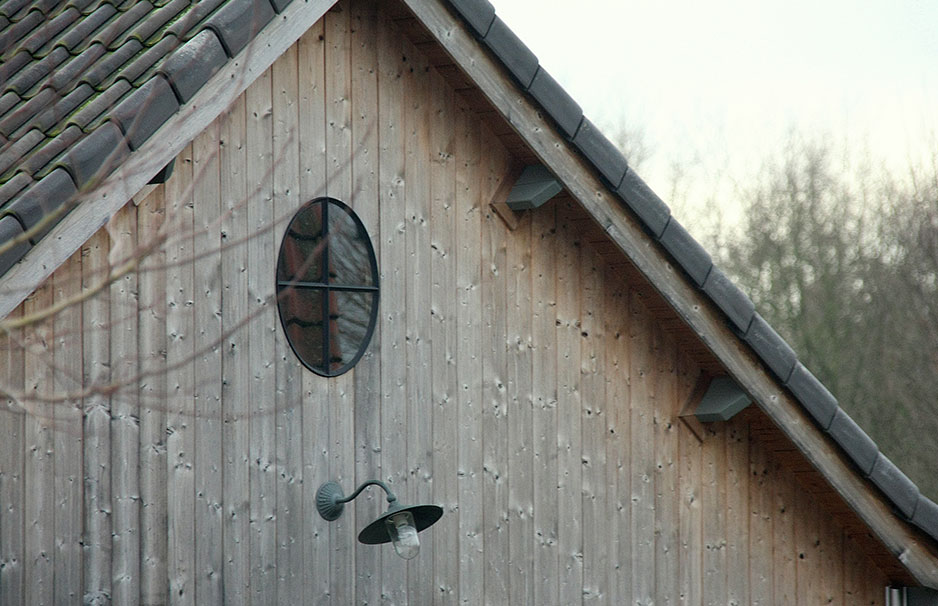 Die Giebelfront eines Holzhauses mit ovalem Fenster und schwarzer Außenlampe darunter, Dachziegel sichtbar