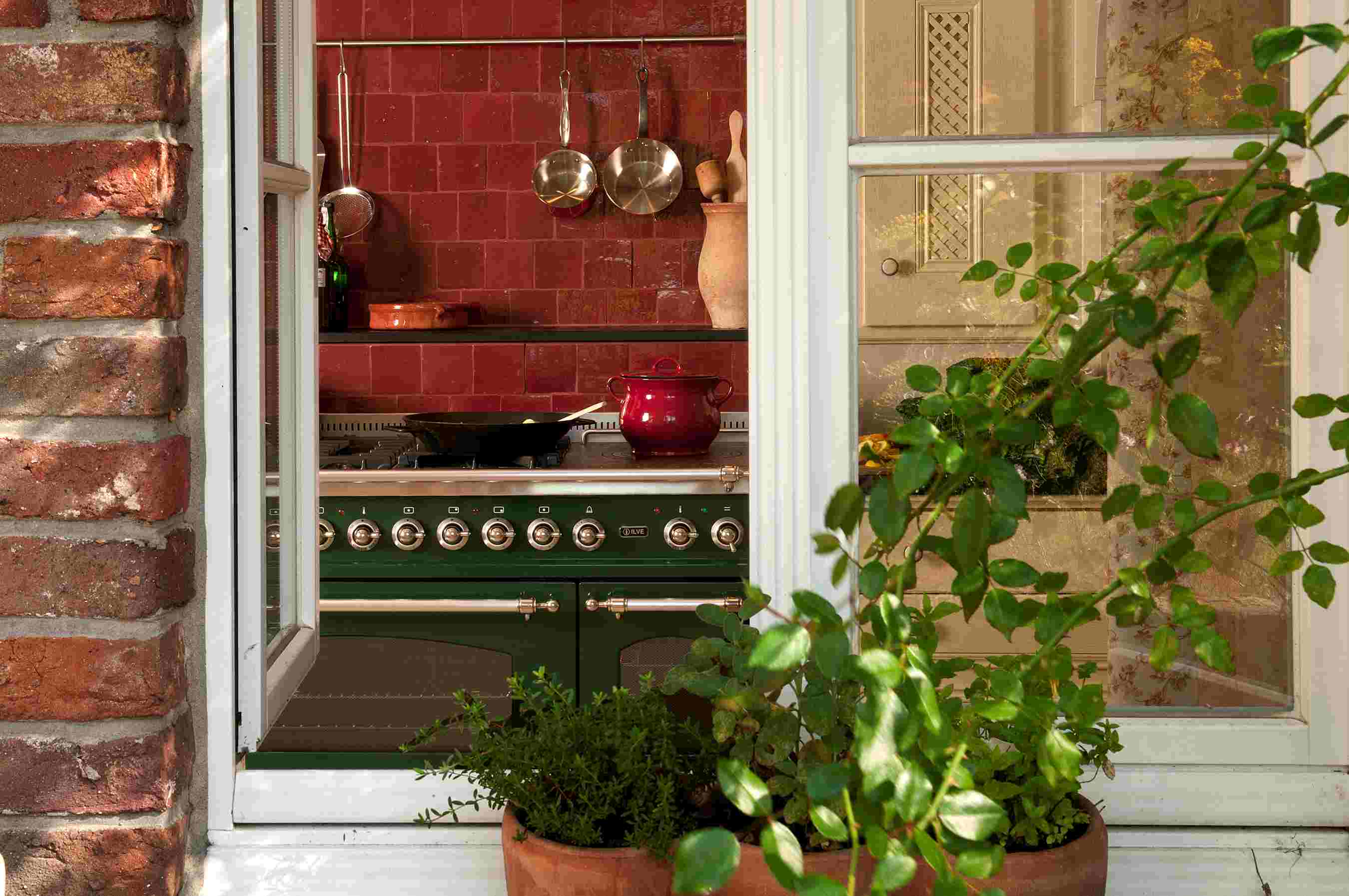 Ein gemütlicher Blick von außen durch ein geöffnetes Fenster auf eine Landhausküche mit grünem Herd, roten Wandfliesen, Töpfen, Küchenutensilien und Kräutern im Blumentopf