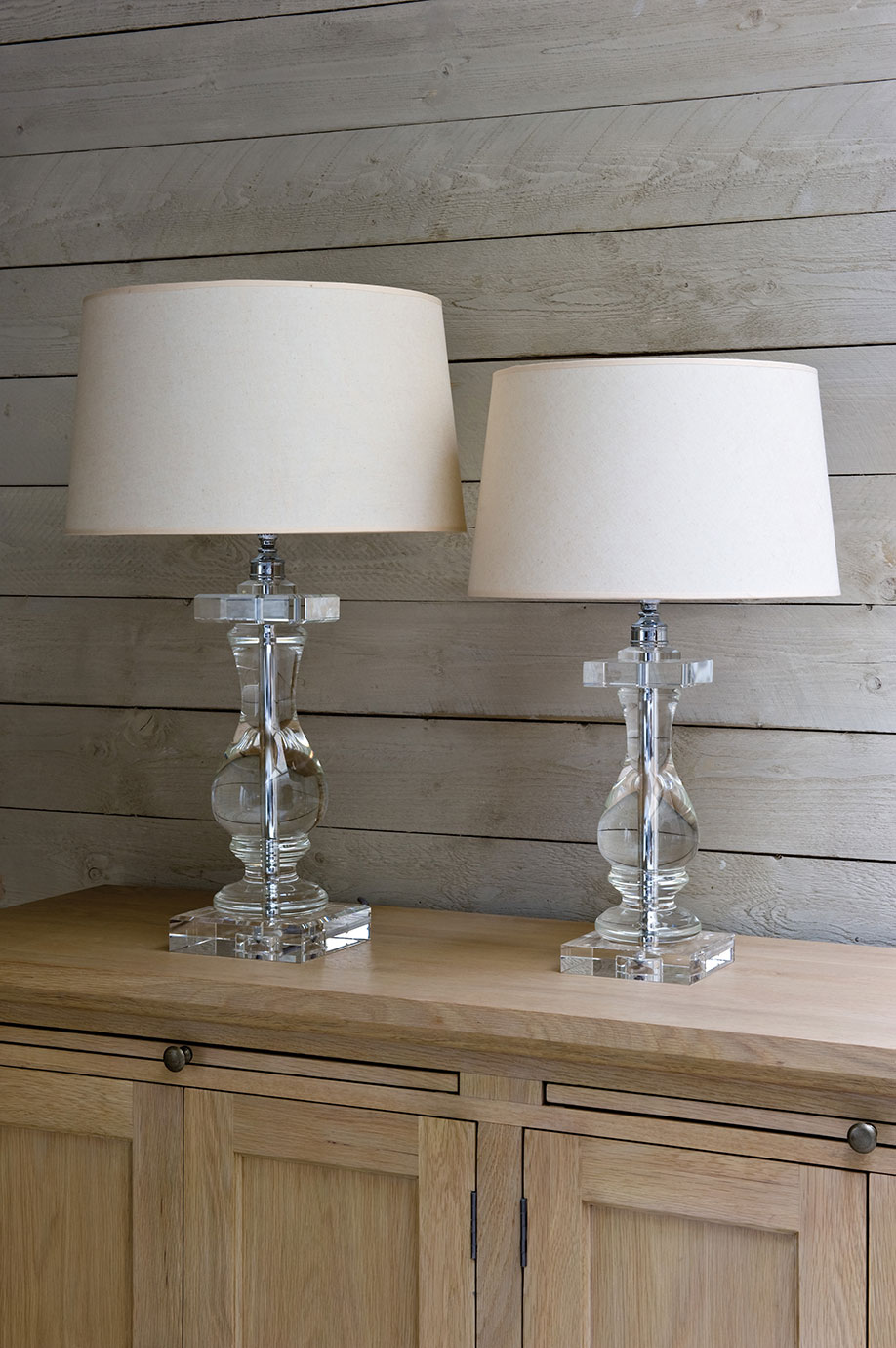 Zwei stilvolle Tischlampen mit transparentem Glasfuß und beigem Lampenschirm auf einer Holzkommode vor einer Holzvertäfelung