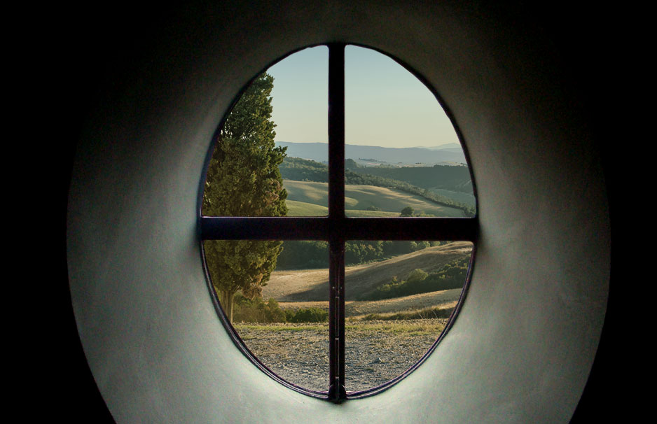 Blick durch ein rundes Fenster mit Kreuzsprossen auf grüne Hügel und Bäume in einer ländlichen Landschaft
