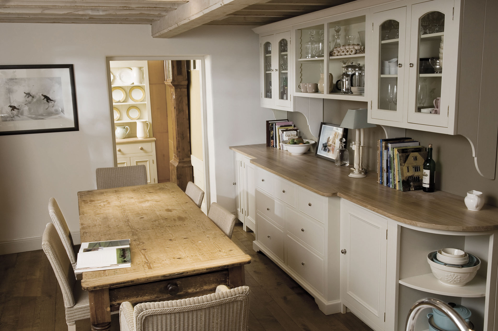 Helles Esszimmer mit rustikalem Holztisch, geflochtenen Stühlen und einem weißen Küchenbuffet im Landhausstil, dekoriert mit Geschirr und Büchern.