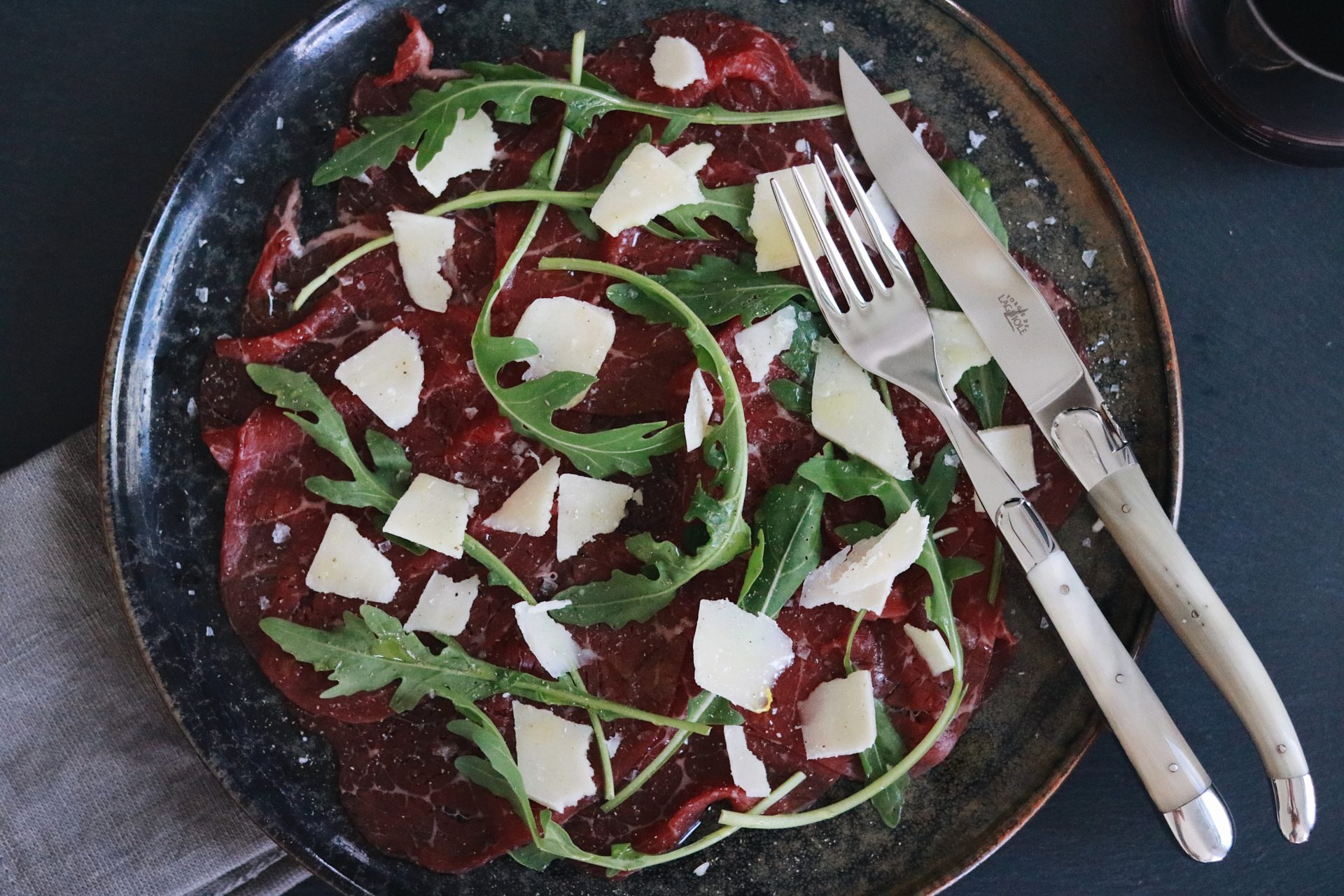 Carpaccio aus Rindfleisch mit frischem Rucola und Parmesanspänen auf einem Teller, daneben Messer und Gabel