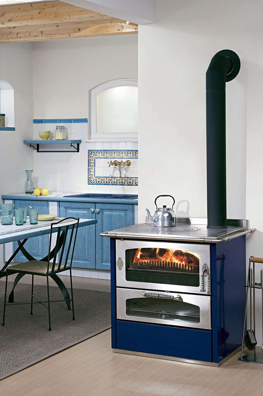 Blauer Küchenofen mit Holzfeuer neben rustikaler Küche mit blauen Schränken und Esstisch