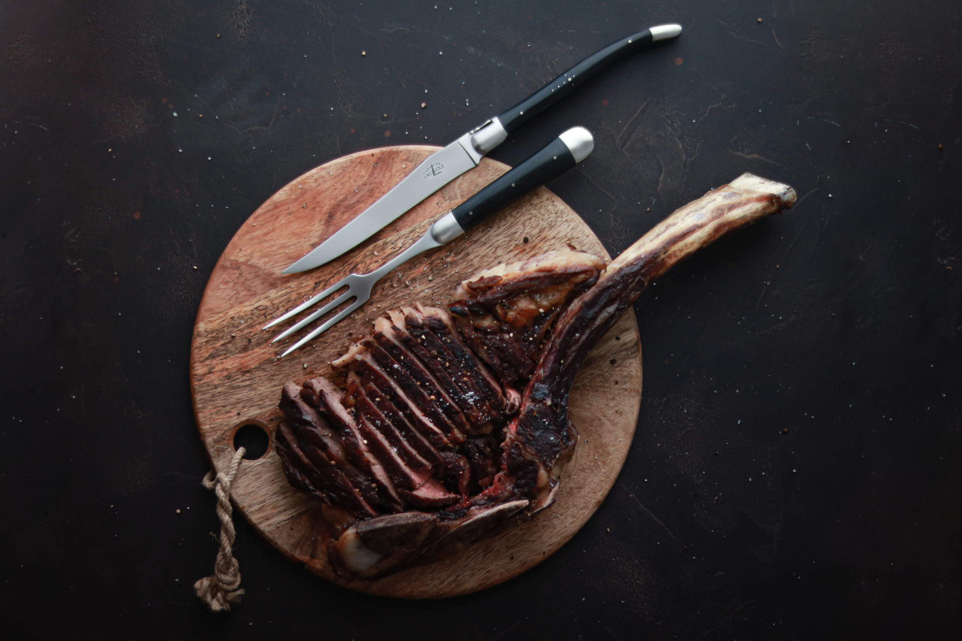 Saftig gegrilltes Tomahawk-Steak auf einem runden Holzbrett mit passendem Besteck, Messer und Fleischgabel