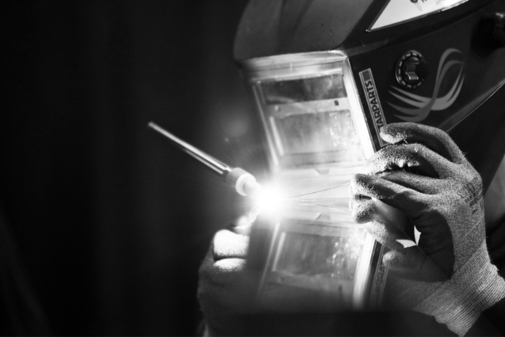 Person mit Schweißhelm und Schutzhandschuhen führt eine Schweißarbeit aus, heller Lichtbogen sichtbar