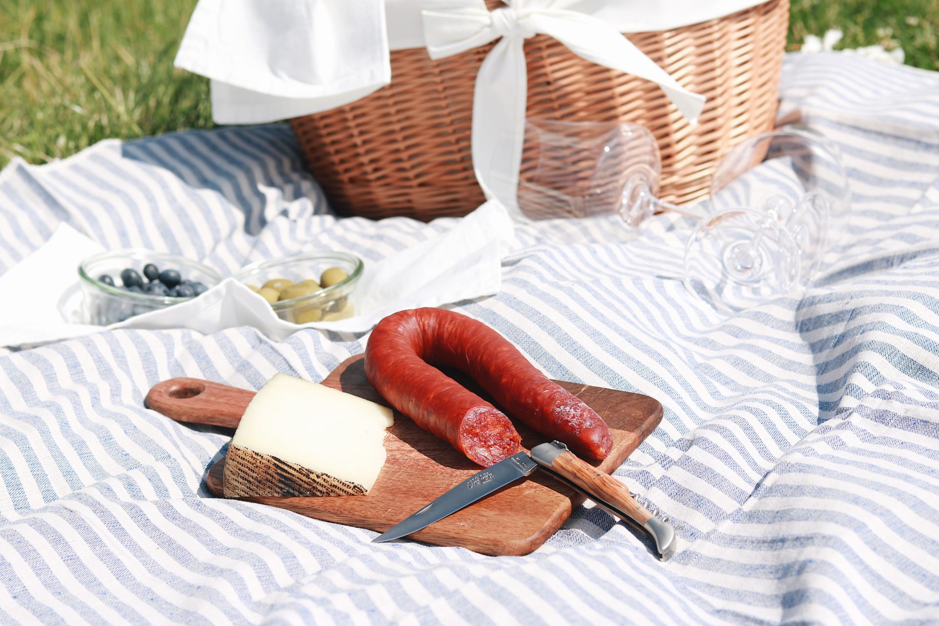 Ein Picknick mit einer Holzplatte mit Wurst, Käse und Messer, daneben Schalen mit Oliven und Blaubeeren auf einem gestreiften Tuch, im Hintergrund ein Weidenkorb.