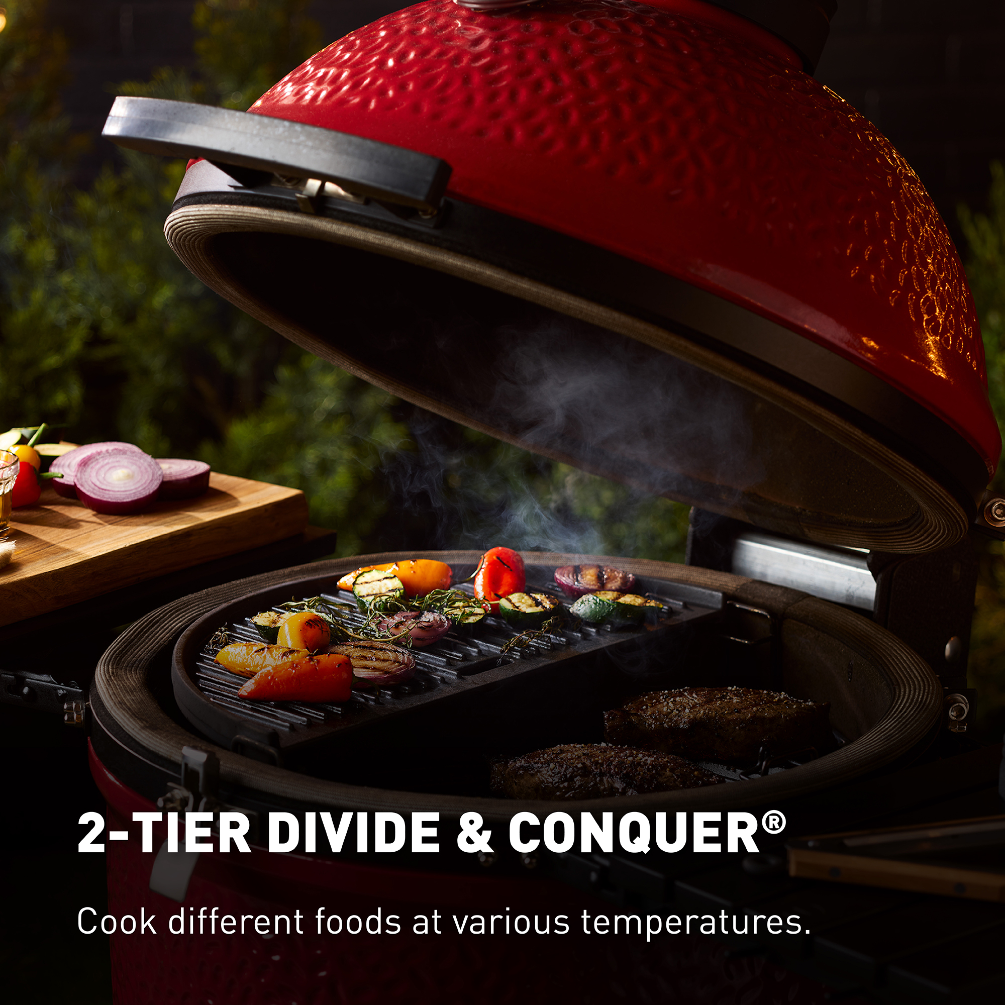 Offener Keramikgrill mit Grillgemüse und Steaks auf zwei Ebenen, daneben ein Holzbrett mit Zwiebeln