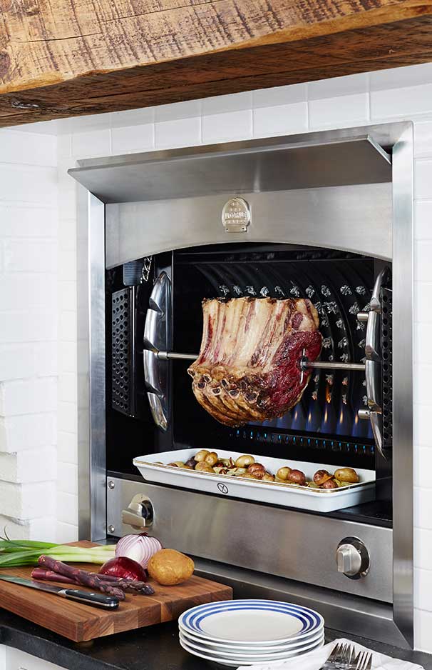 Ein moderner Einbaugrill aus Edelstahl mit einem großen Stück Fleisch am Drehspieß. Darunter steht eine weiße Auflaufform mit kleinen Kartoffeln und Gemüse. Im Vordergrund ein Schneidebrett mit Zutaten sowie gestapelte Teller.