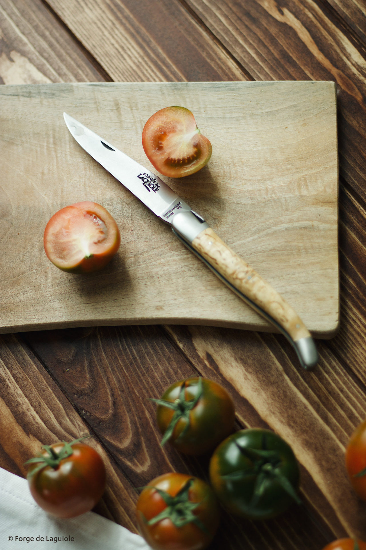 Ein Laguiole Messer mit hellem Holzgriff liegt auf einem Schneidebrett neben halbierten und ganzen Tomaten. Perfekt für die Zubereitung in der Küche.