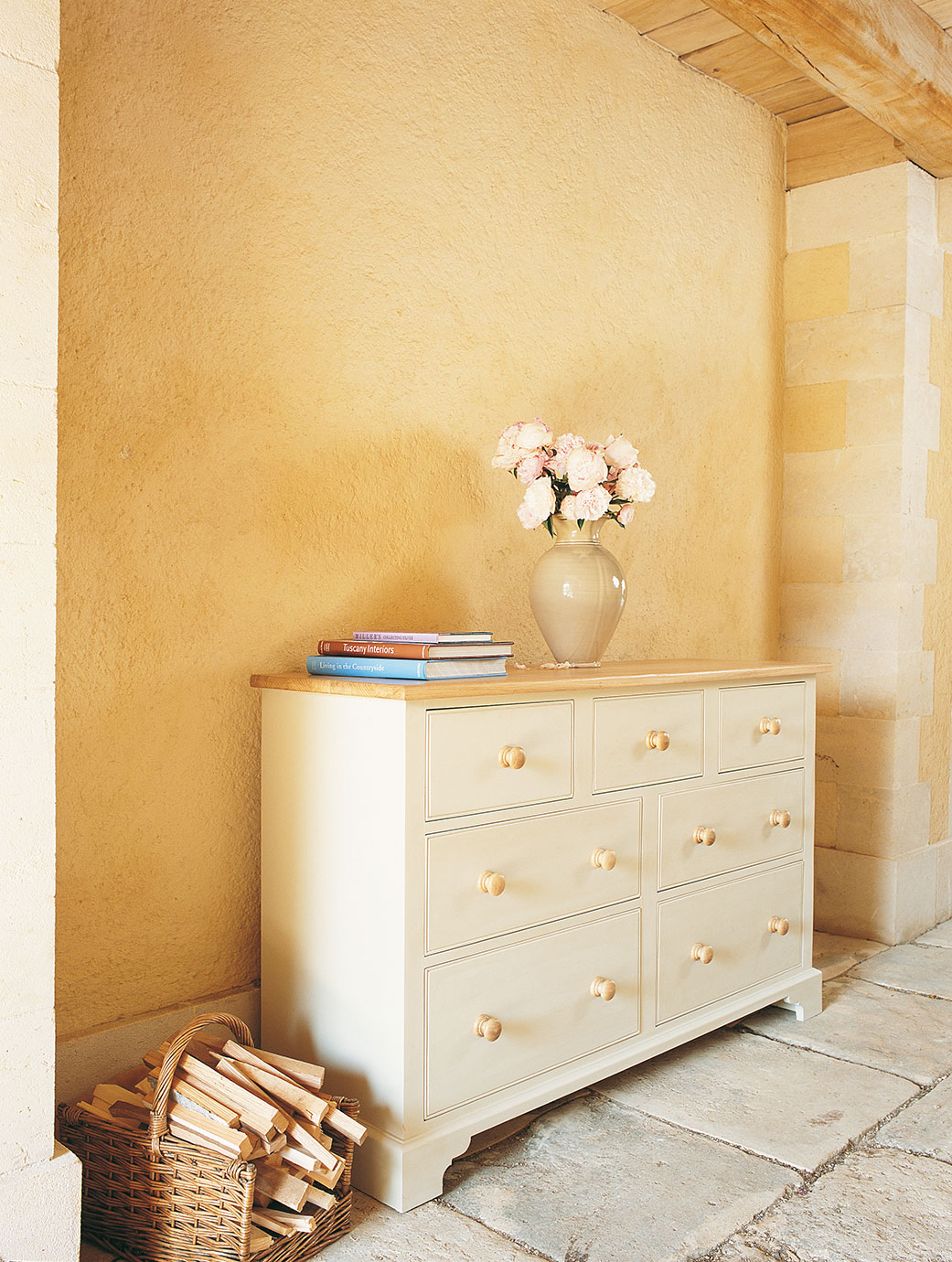 Landhaus Kommode in Weiß mit sieben Schubladen, Holzgriffen, dekorativer Vase mit Blumen und Büchern obenauf, Holzkorb daneben