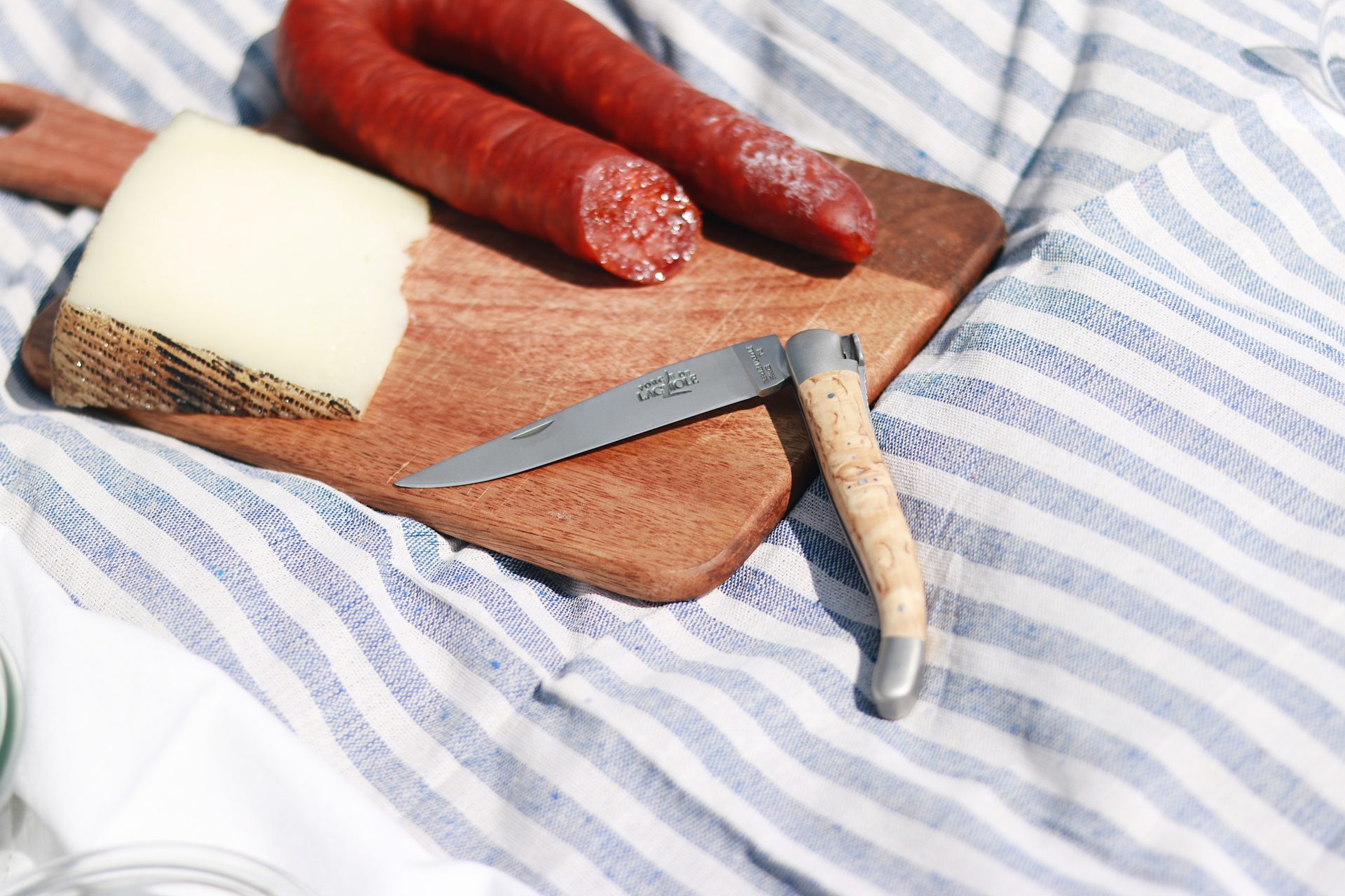 Laguiole Taschenmesser mit Holzgriff neben einem Stück Hartkäse und einer Chorizo auf einem Holzbrett, liegend auf gestreiftem Tuch