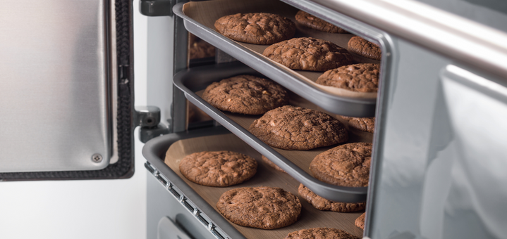Drei Backbleche mit Schokoladenkeksen werden in einem geöffneten Ofen gebacken.