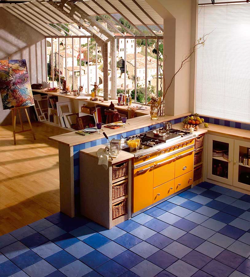 Moderne Wohnküche mit gelbem Herd, blau karierten Fliesen, viel Tageslicht und angrenzendem Atelierbereich mit Staffelei und Gemälden