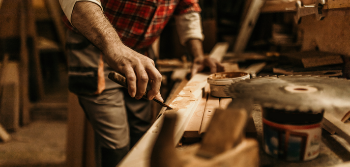 Hand eines Mannes in kariertem Hemd, der in einer Werkstatt ein Stück Holz mit einem Hobel bearbeitet, im Vordergrund verschiedene Werkzeuge