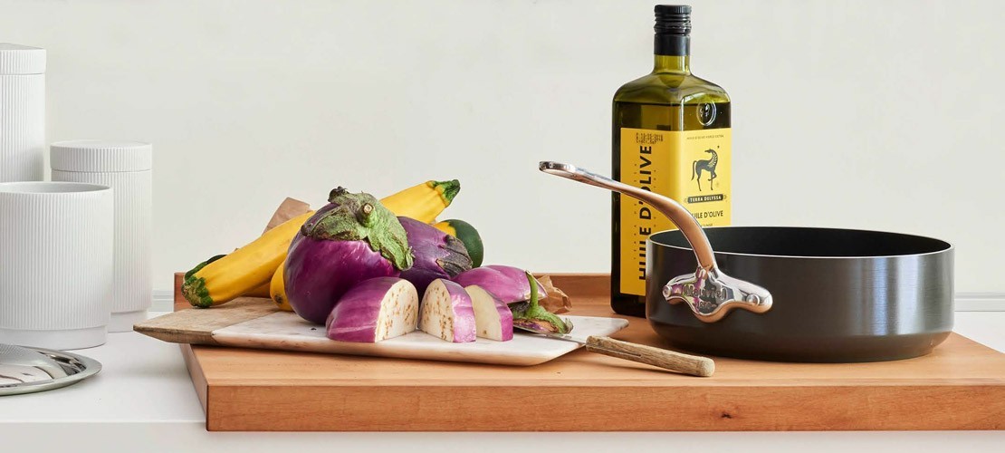Auf einem Holztisch stehen ein Schneidebrett mit geschnittenen Auberginen und Zucchini, eine schwarze Pfanne mit Metallgriff sowie eine Flasche Olivenöl vor weißen Vorratsdosen.