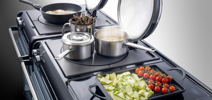 Kochfeld mit Edelstahl-Töpfen, Pfanne mit Spargel, kochendem Wasser und einer Grillplatte mit Zucchini und Cocktailtomaten