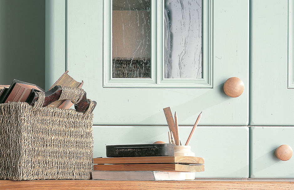 Geflochtener Korb mit alten Büchern, Holzstiftehalter und Bücherstapel auf Holzoberfläche vor mintgrünem Schrank mit Glastüren