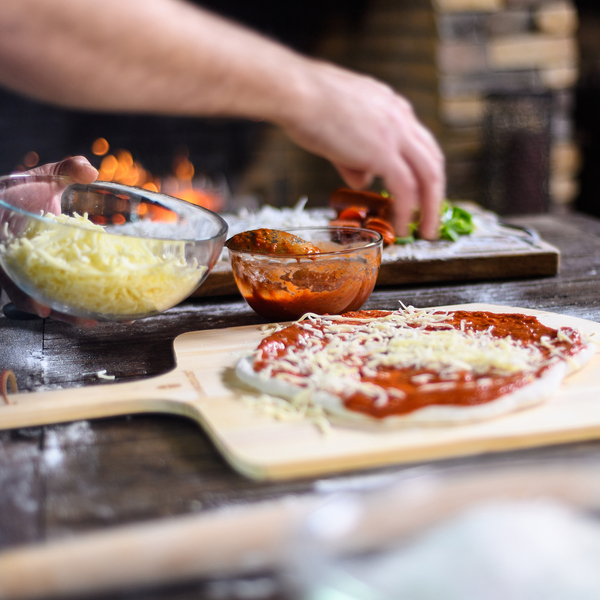 Eine Hand verteilt geriebenen Käse auf einen mit Tomatensauce bestrichenen Pizzateig, daneben eine Schüssel mit Pizzasauce, im Hintergrund brennt ein Holzofen.