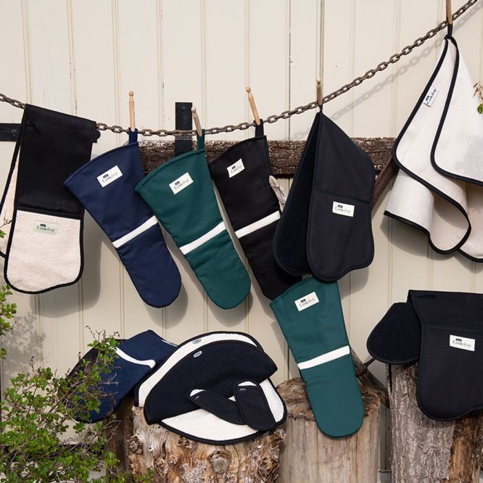 Verschiedene Ofenhandschuhe und Topflappen aus Stoff in Grün, Blau, Schwarz und Cremefarben hängen an einer Kette im Freien vor einem Holzzaun.
