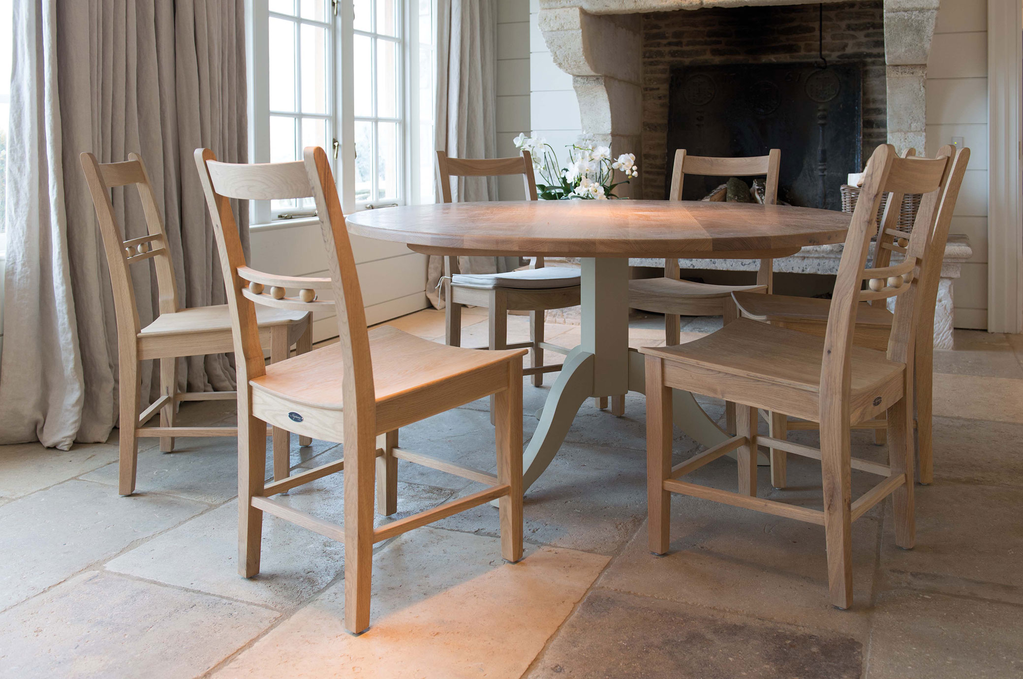 Heller Holztisch rund mit sechs passenden Holzstühlen, dekoriert mit weißen Blumen, in einem lichtdurchfluteten Esszimmer mit Steinkamin