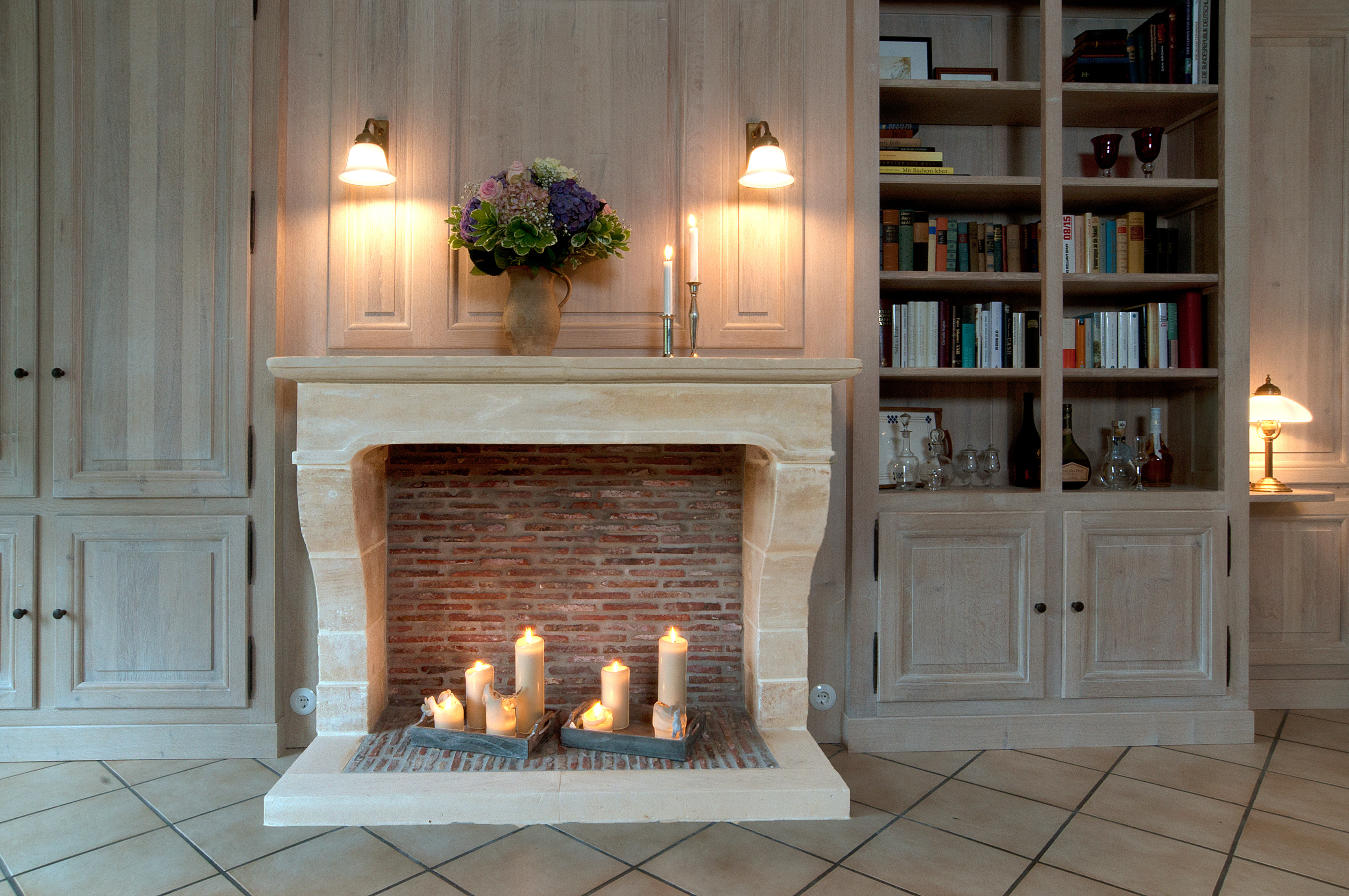 Wohnzimmer mit Kamin, in dem mehrere Kerzen brennen. Über dem Kamin stehen ein Blumenstrauß in einer Vase und zwei Kerzenhalter. Rechts ein Bücherregal, links Holzschränke, warme Beleuchtung.