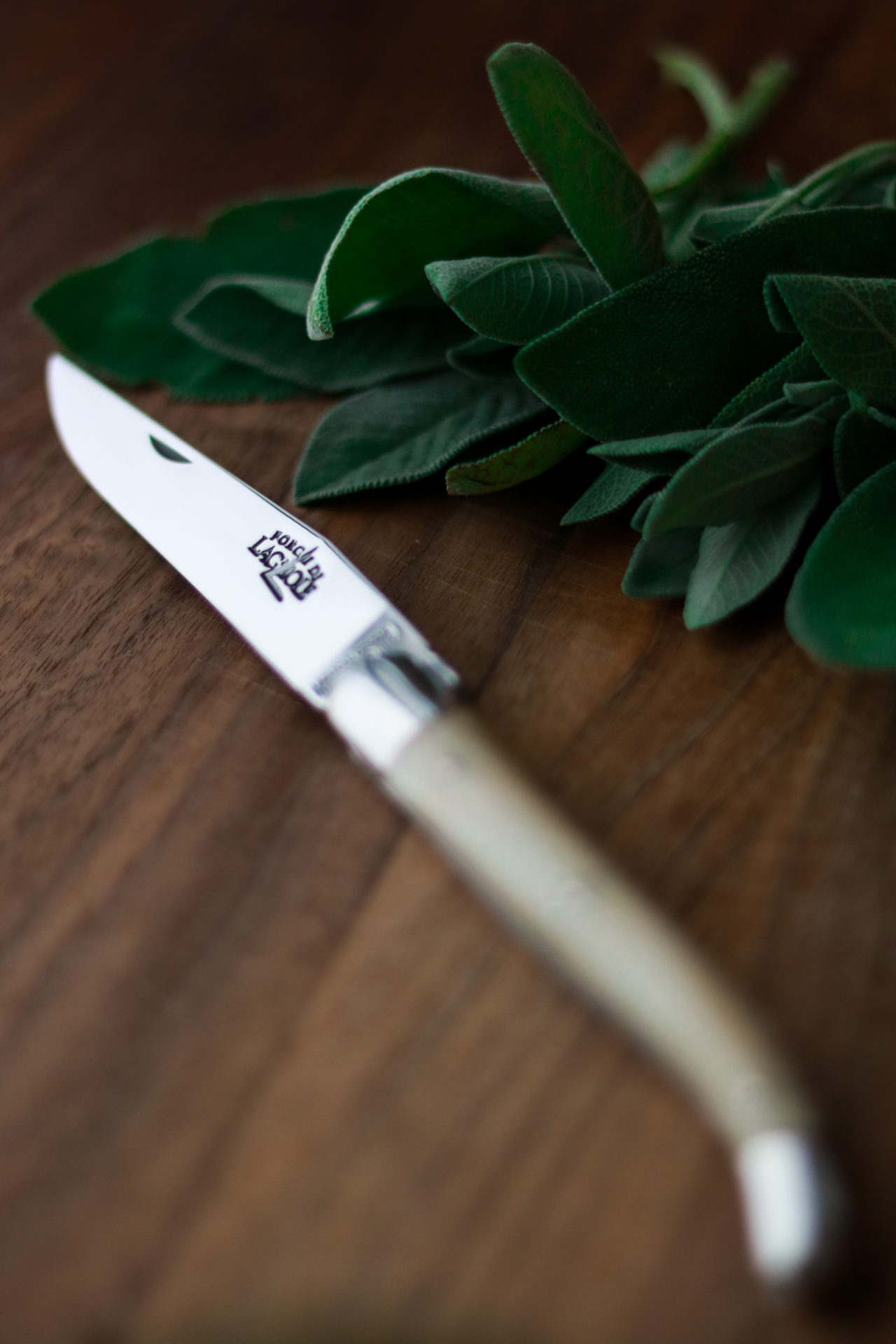 Laguiole Messer mit hellem Griff, liegend auf einem Holzbrett neben frischen Salbeiblättern