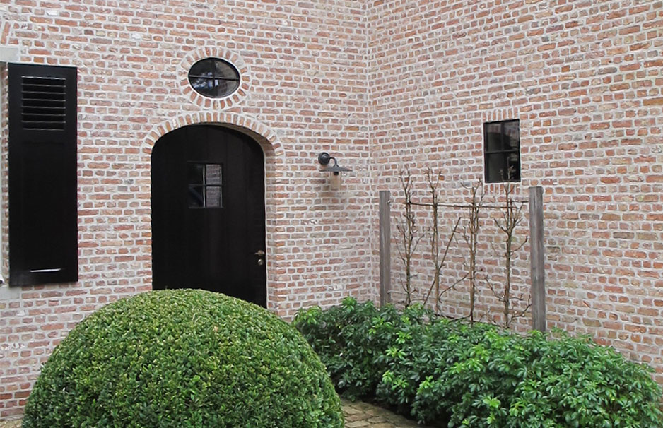 Backsteinfassade mit schwarzer Eingangstür, rundem Fenster über der Tür, blickdichtem Fensterladen und einem kugelförmigen Buchsbaum im Vorgarten