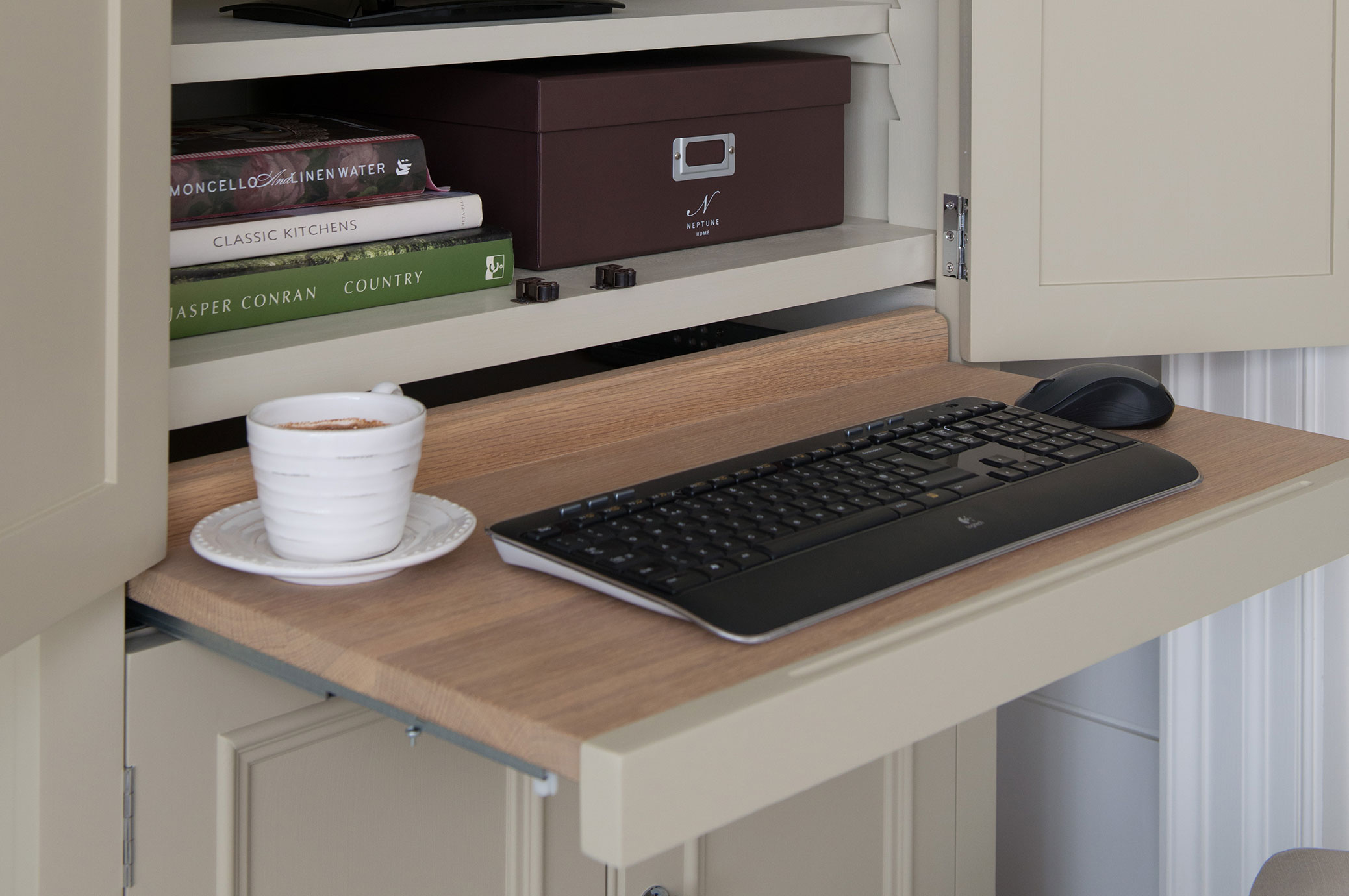 Arbeitsplatz im Schrank mit ausziehbarer Holzplatte, schwarzer Tastatur, Maus und weißer Kaffeetasse, im Hintergrund Bücher und Aufbewahrungsboxen