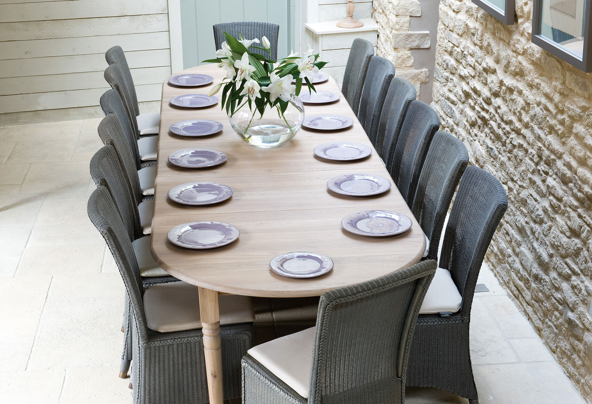 Langer Holztisch mit geflochtenen Stühlen, lila Tellern und Vase mit weißen Lilien in hellem Esszimmer