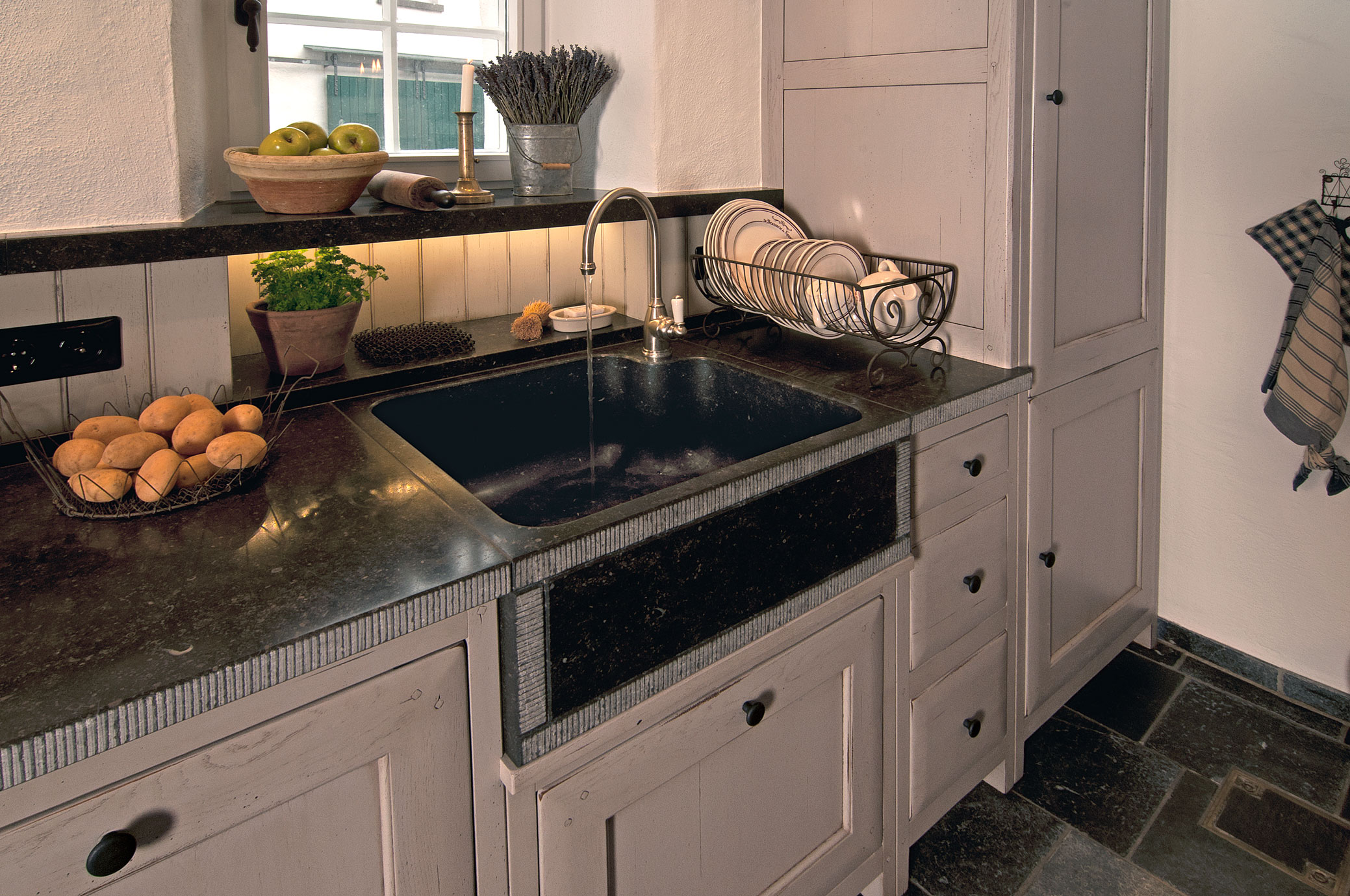 Küchenspüle in Landhausstil mit schwarzer Natursteinplatte, Ablage mit frischen Äpfeln, Kartoffeln, Geschirrkorb und Kräutertopf