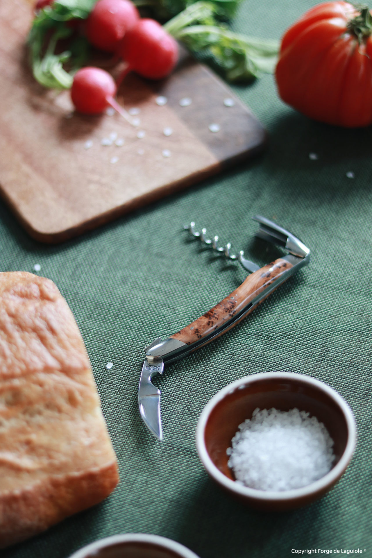 Edler Laguiole Korkenzieher mit Holzgriff liegt auf grünem Tischtuch, daneben frische Zutaten wie Brot, grobes Salz, Radieschen und Tomate