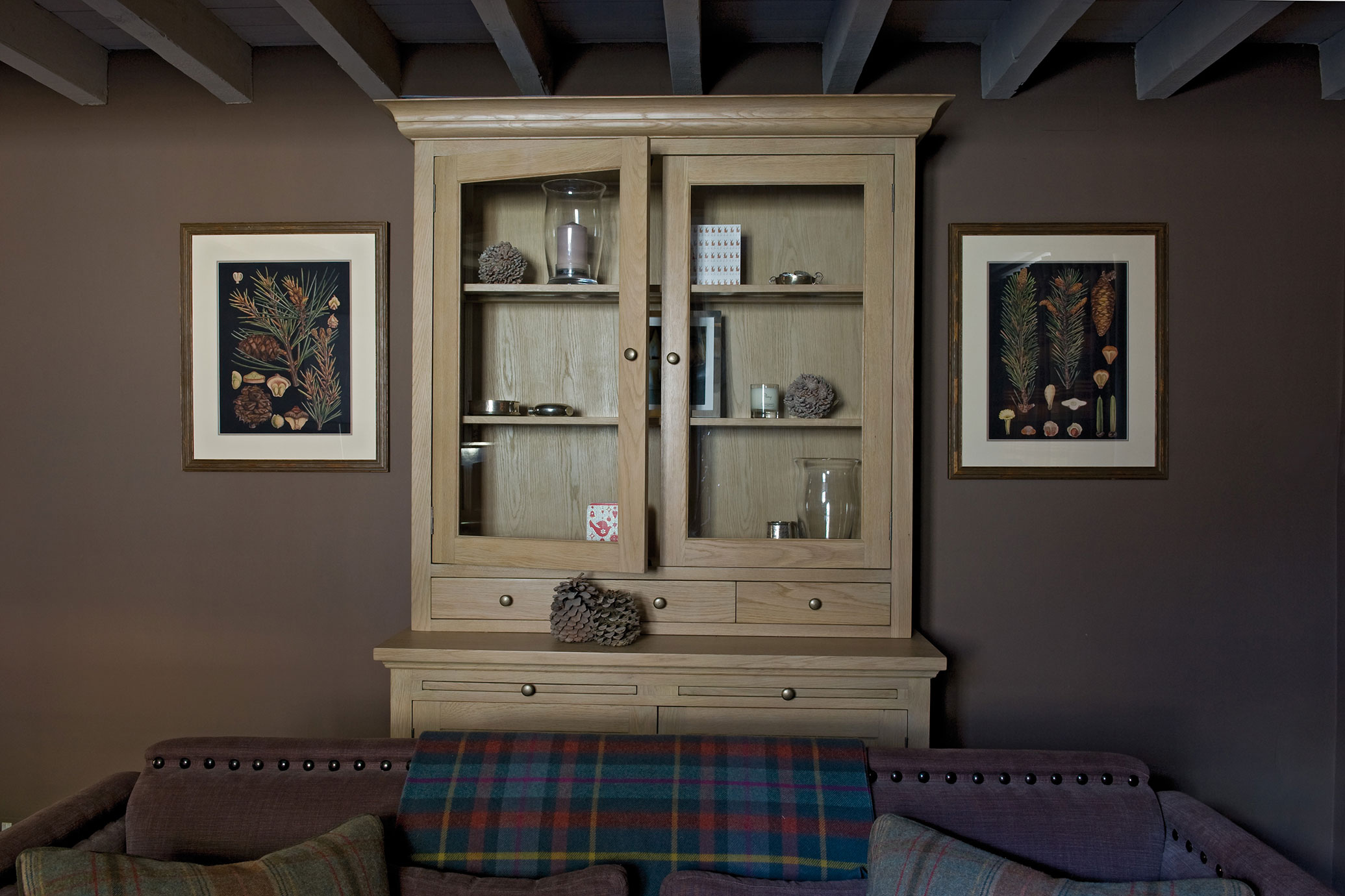 Gemütliches Wohnzimmer mit einem großen Holzvitrinenschrank, botanischen Wandbildern an den Wänden und einem Polstersofa mit karierter Decke im Vordergrund