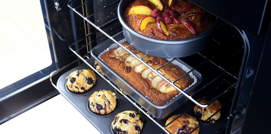 Offener Backofen mit einem Obstkuchen in einer runden Form, einem rechteckigen Apfelkuchen und vier Blaubeermuffins auf dem Blech
