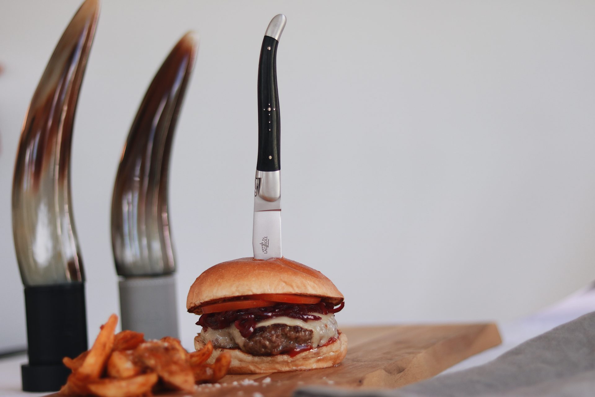 Saftiger Burger mit Tomate, Käse und Zwiebeln, in dessen Mitte ein Messer steckt, dazu knusprige Pommes auf einem Holzbrett serviert