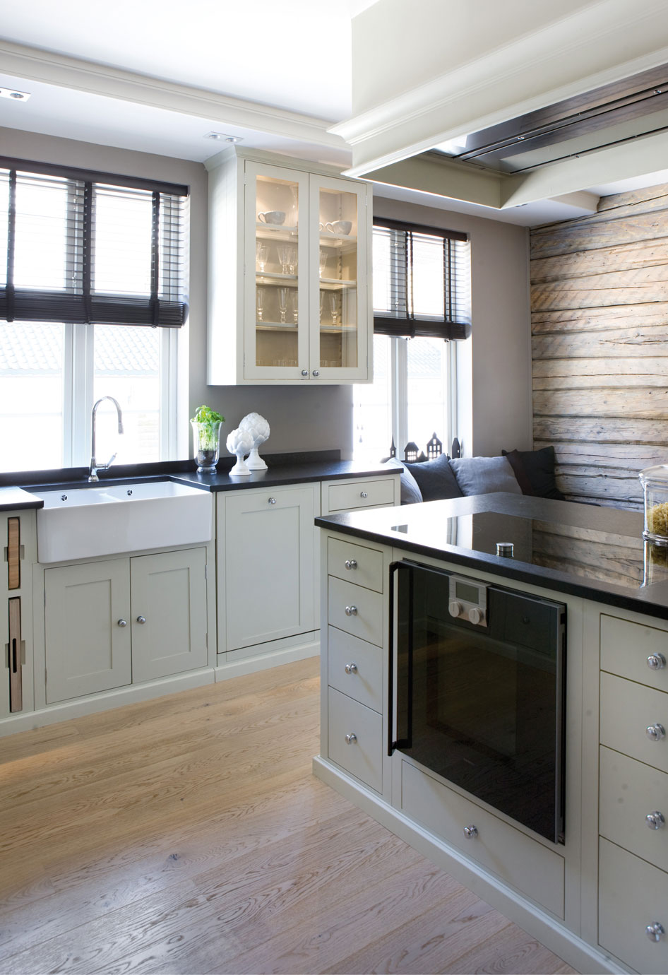 Helle, moderne Landhausküche mit weißen Schränken, schwarzen Arbeitsplatten, eingebautem Backofen, Spülbecken vor Fenstern und Sitzbank an Holzwand