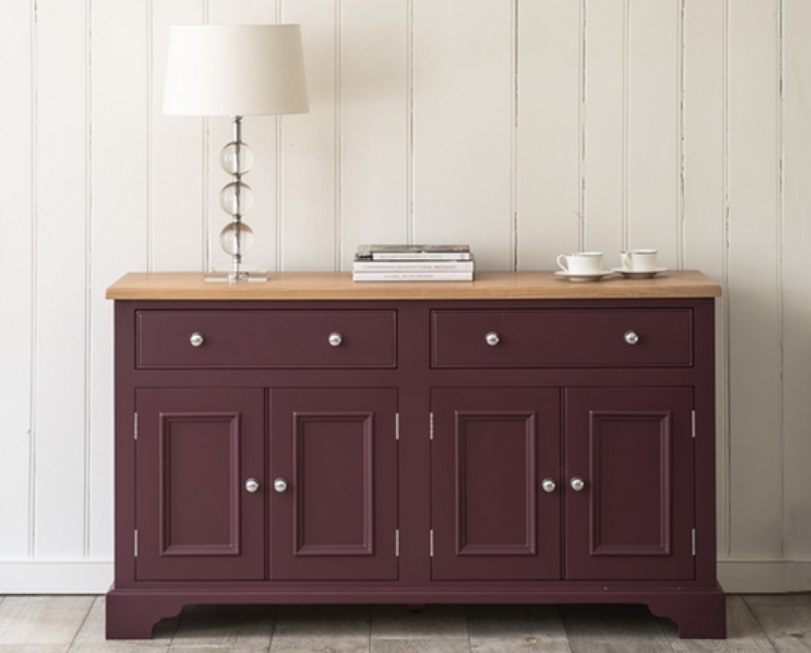 Elegantes Sideboard in Dunkelrot mit vier Türen und zwei Schubladen, oben mit Lampe, Büchern und Tassen dekoriert, steht vor weißer Holzvertäfelung