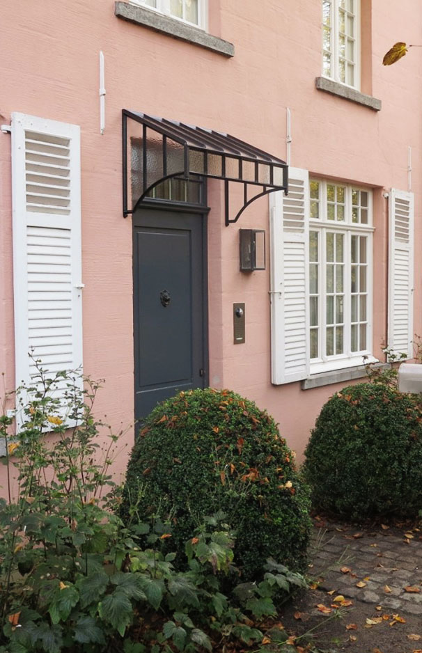 Schwarze Haustür mit Glaskuppel-Vordach, daneben weiße Fensterläden und große grüne Buchskugeln vor einem rosa Haus