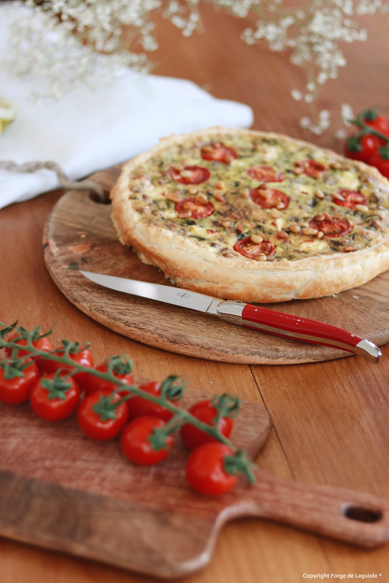 Eine Gemüsequiche mit Kirschtomaten auf einem runden Holzbrett, daneben ein Messer mit rotem Griff und eine Rispe frischer Kirschtomaten auf einem kleinen Holzbrettchen