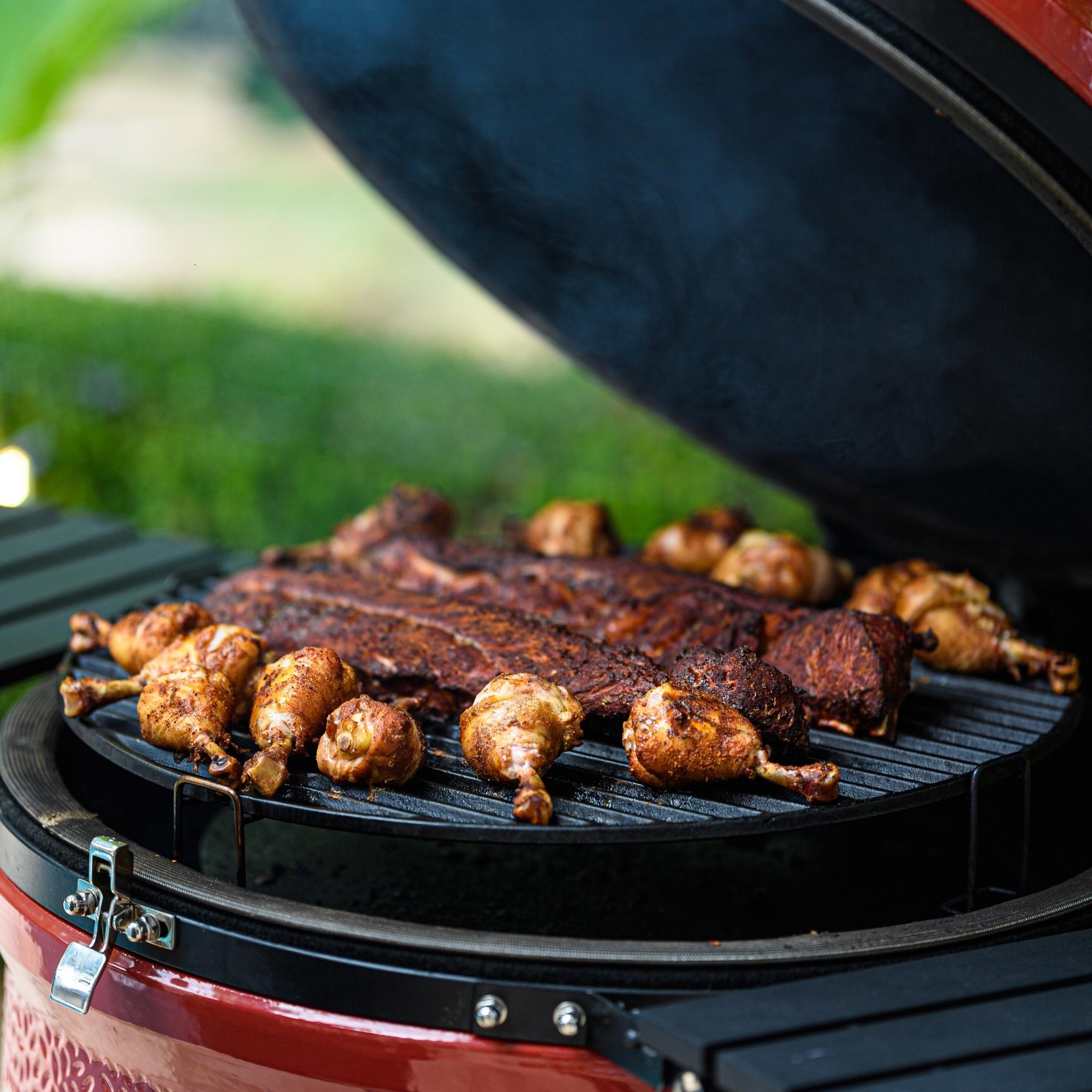 Geöffneter runder Keramikgrill mit gegrillten Hähnchenschenkeln und Spareribs, Grill steht im Freien auf einer Wiese