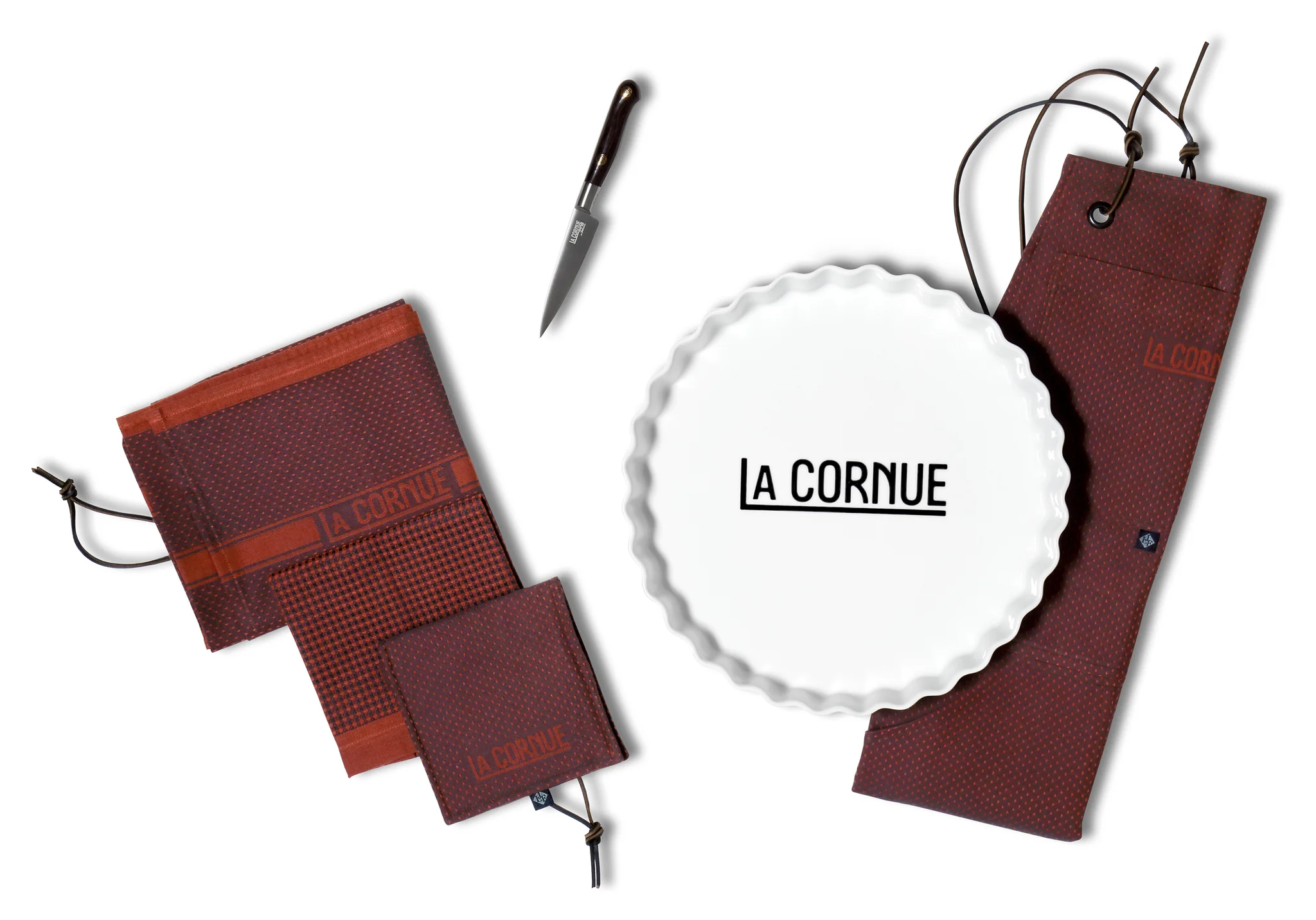 La Cornue Backset bestehend aus einer weißen Keramik-Tarteform mit La Cornue Logo, einem Messer und braunen Küchentextilien mit Kordelzug