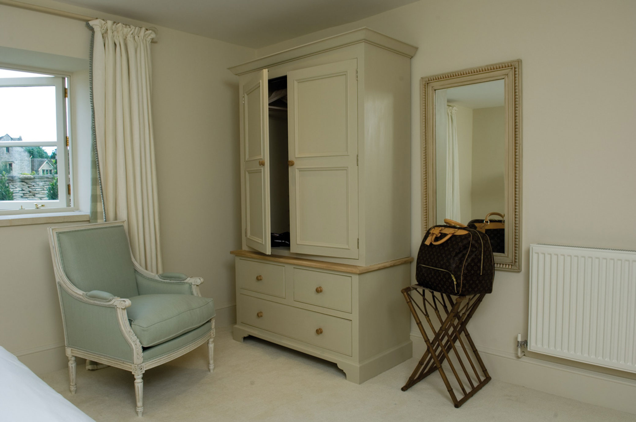 Schlafzimmer mit hellgrünem Sessel, cremefarbenem Kleiderschrank, großem Wandspiegel und einer Reisetasche auf einem Gepäckständer