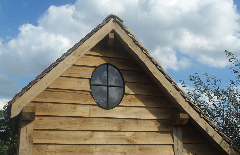 Vorderseite eines Holzhauses mit rundem, verglastem Fenster, Dachziegeln und bewölktem Himmel im Hintergrund