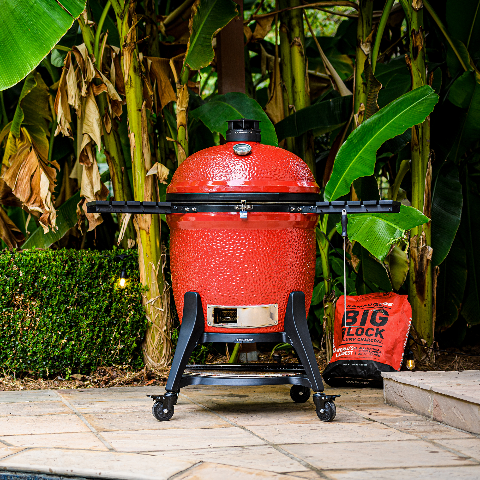 Roter Kamado Joe Classic II Keramikgrill auf Rollwagen mit Ablagen, im Garten neben einem Sack Holzkohle