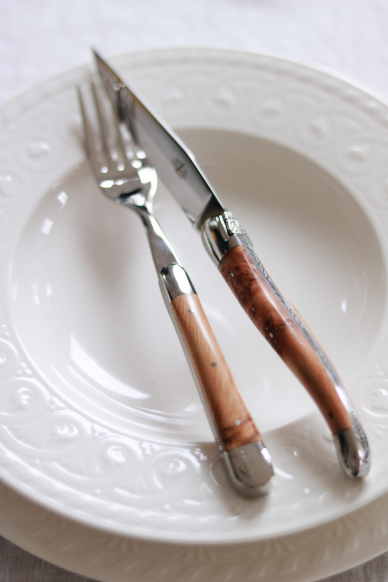 Gabel und Messer mit Holzgriffen liegen gekreuzt auf einem weißen, verzierten Teller aus Porzellan