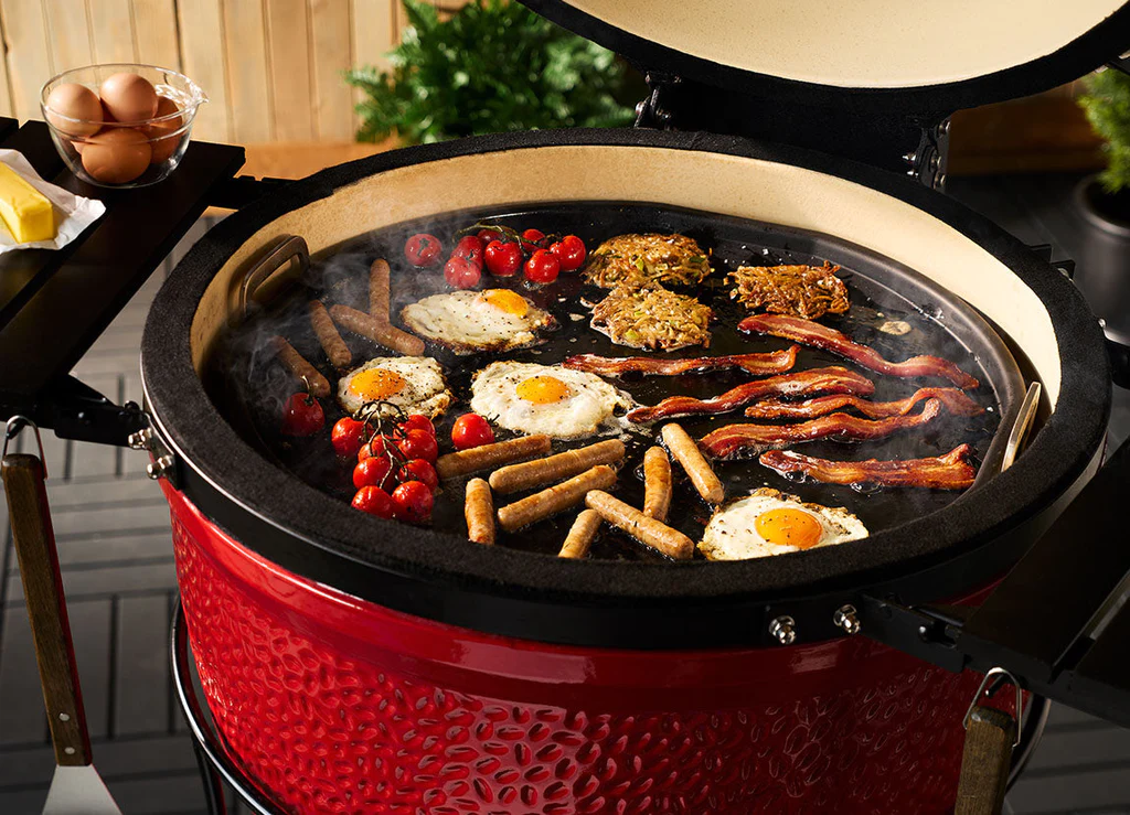 Frühstück wird auf einem roten Keramikgrill gegrillt: Spiegeleier, Speck, Würstchen, Rösti und Tomaten auf Grillplatte