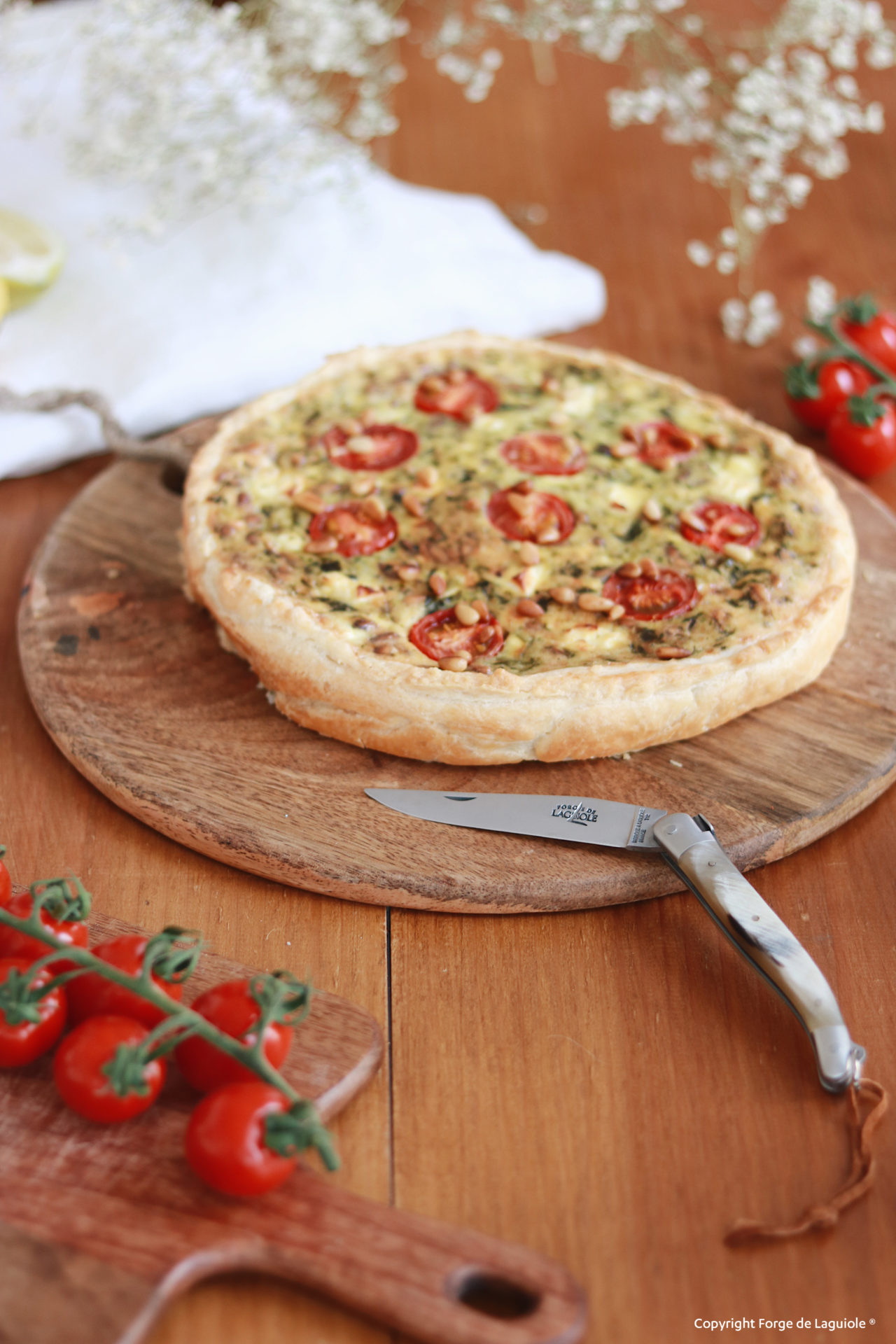 Quiche mit Tomaten und Käse auf rundem Holzbrett, daneben ein Laguiole-Messer und frische Cherrytomaten